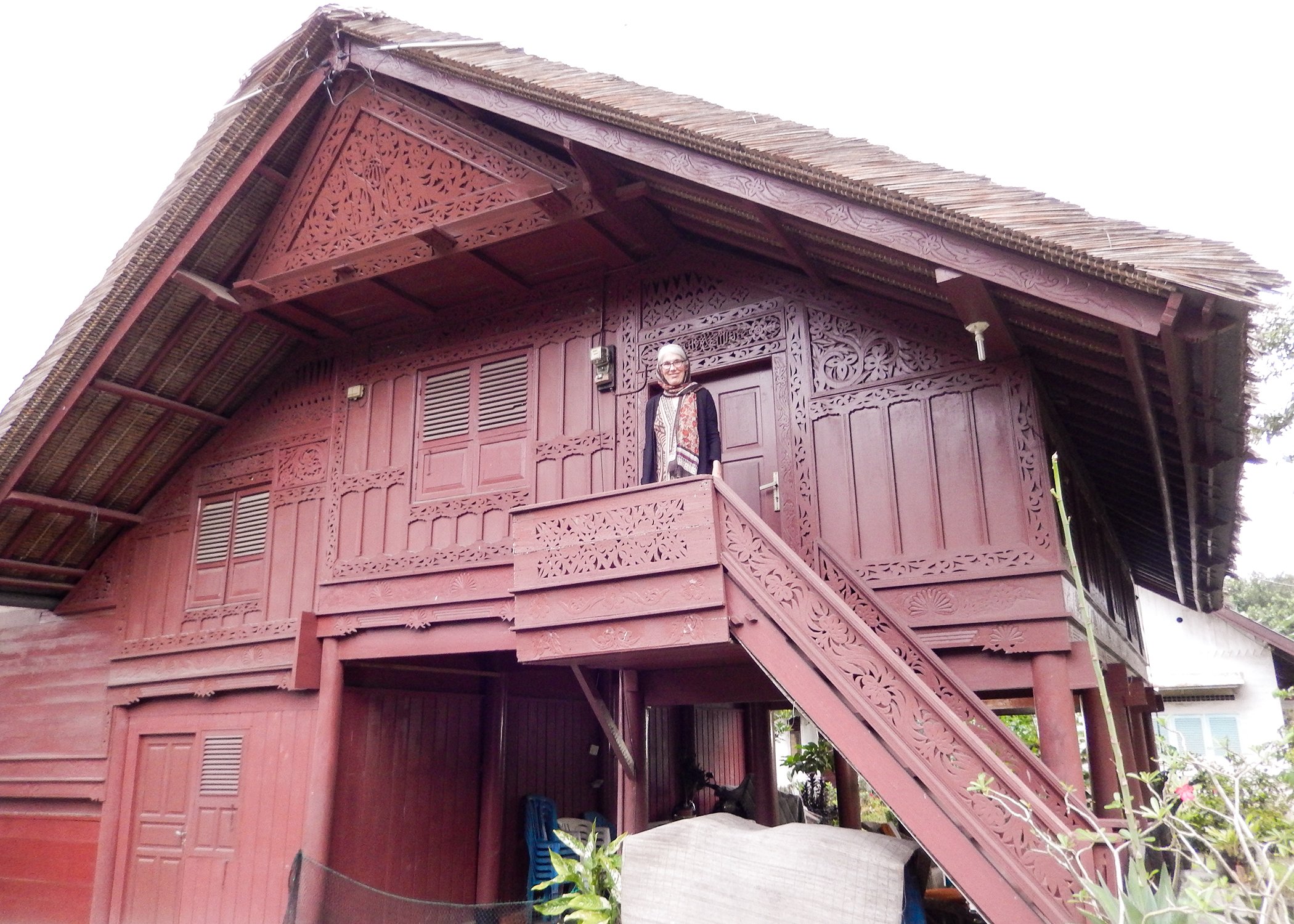 Image of a woman standing on the steps of a traditional Aceh house